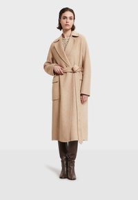 Abrigo de mezcla de lana beige con cintura ceñida, cuello de solapa, dos bolsillos frontales y mangas largas, llevado sobre una blusa con estampado y botas marrones hasta la rodilla.