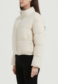 La doudoune couleur crème présente un col haut, une fermeture éclair à l'avant et des poches latérales, avec un design court et une texture légèrement matelassée.