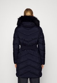 Námořnický péřový kabát s odepínací kapucí z umělé kožešiny. Má pás v pase a vzor prošívání ve tvaru rybí kosti na odolném vnějším materiálu.