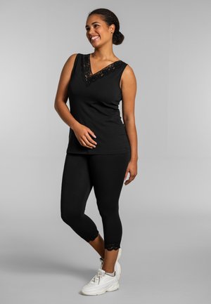 Femeie zâmbitoare, purtând un top negru fără mâneci cu dantelă și colanți scurți, cu adidași albi, stând pe un fundal gri.