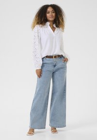 White shirt with lace long sleeves, v-neck design. Light blue wide-leg jeans, brown belt, and white heeled sandals. Simple, casual outfit.
