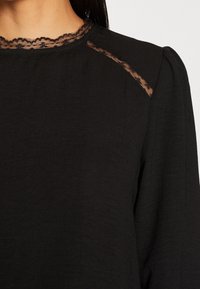 Haut noir à manches longues avec bordure en dentelle le long du décolleté et de l'épaule, tissu texturé, porté par une personne aux cheveux foncés.