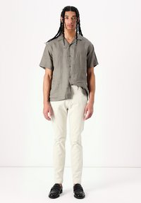 Camicia grigia in lino a maniche corte con chiusura a bottoni, abbinata a pantaloni aderenti di colore chiaro e mocassini neri. Tessuto semplice e strutturato.
