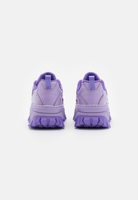 Ett par kraftiga lila sneakers visade från baksidan, med tjocka texturerade sulor och dragremmar på en vit bakgrund.