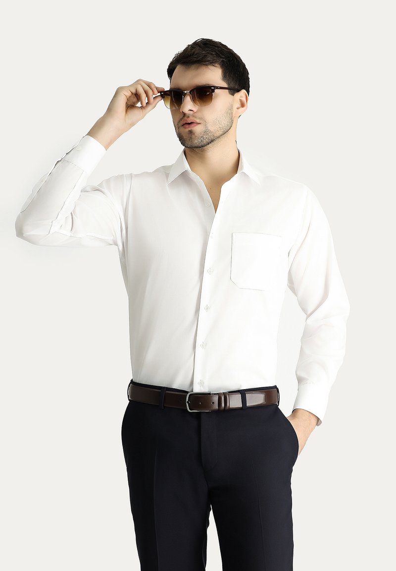 Camicia bianca a maniche lunghe con colletto button-down e taschino, abbinata a pantaloni scuri e cintura in pelle marrone, accessoriata con occhiali da sole.