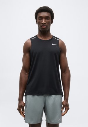 Άνδρας που στέκεται με το πρόσωπο προς τα εμπρός, φοράει μαύρο αμάνικο αθλητικό τοπ και γκρι σορτς σε απλό ανοιχτό φόντο.