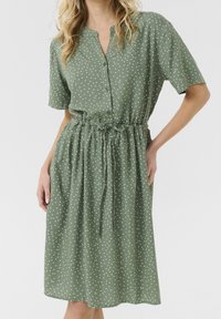 Grön klänning med polka prickar, knäppning framtill, korta ärmar, elastisk midja och snöre. Tillverkad av ett lätt material med en avslappnad passform.
