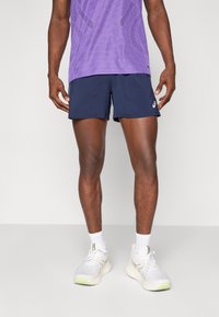 Sportliches Herrenoberteil in Lila mit Muster, marineblaue Shorts, weiße Sneaker mit grünen Akzenten und weiße Socken. Sportlicher Schnitt und glatte Textur.