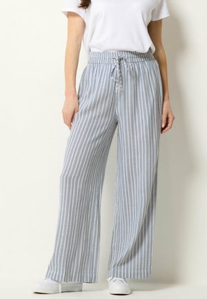 Kobieta ubrana w jasnoniebieskie szerokie spodnie w paski z troczkiem w talii i białe trampki, w połączeniu z gładką białą koszulką z krótkim rękawem.