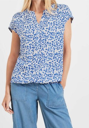 Femme portant un chemisier blanc à manches courtes avec un motif de feuilles bleues et un pantalon large bleu clair avec de grandes poches avant.