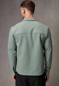 Camisa verde de manga larga con cuello, textura suave, puños abotonados y una costura horizontal en la espalda.