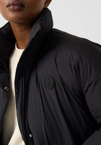 Zwarte gewatteerde jas met een hoge kraag, voorzien van een ronde logopatch op de borst. Gladde textuur en ritssluiting.