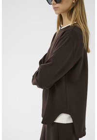 Sudadera marrón con un corte relajado, dobladillo redondeado y mangas largas; llevada sobre una camiseta blanca; la tela parece suave y lisa.