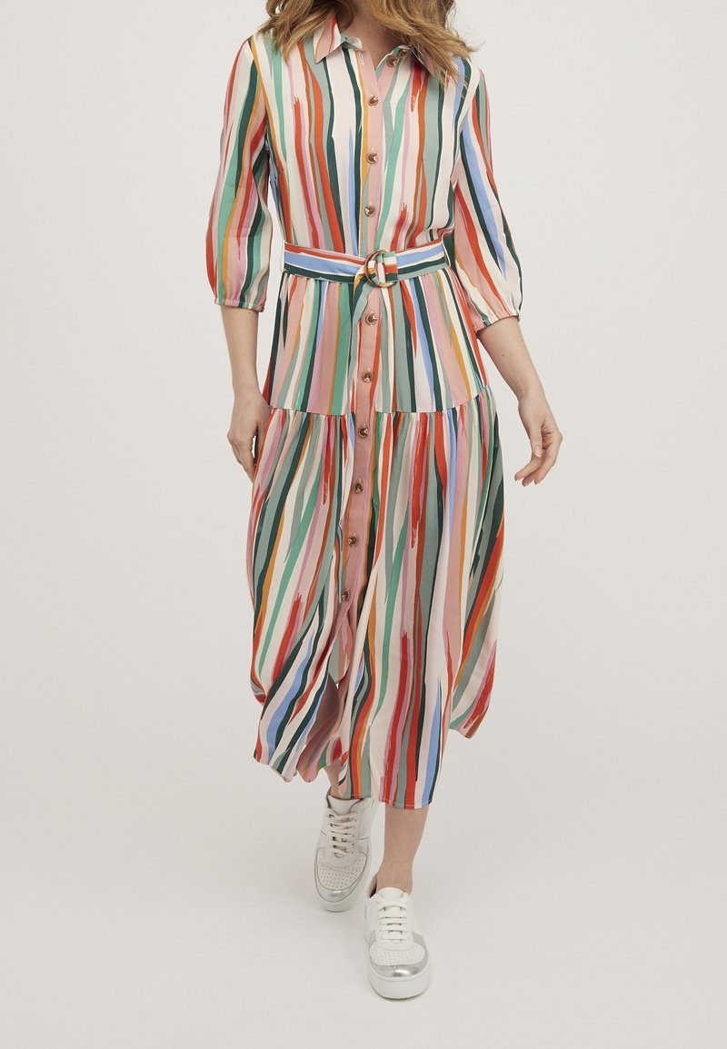 Vrouw draagt een multicolore midi-jurk met verticale strepen, knopen en een riem, in combinatie met witte sneakers, terwijl ze vooruit loopt.