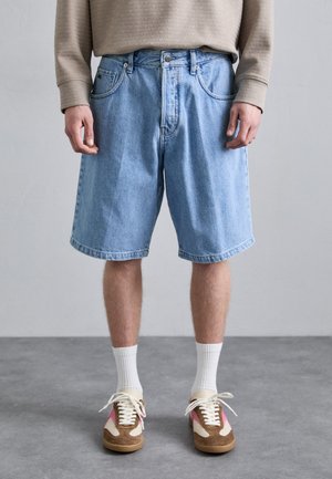 Shorts en denim bleu clair avec une coupe décontractée, comportant des poches avant et une fermeture à bouton. Portés avec des chaussettes blanches et des baskets marron.