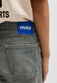 Grå denimbyxor med en blå etikett som visar "HUGO" på baksidan. Funktioner inkluderar en femfickorsdesign och kontrasterande sömmar.