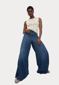 Brede denim jeans in een donkerblauwe was, gecombineerd met een crèmekleurige gebreide mouwloze top met een ruitpatroon.