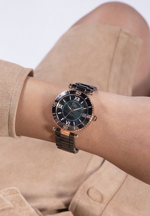 Håndled iført et analogt ur i sort og guld med romertal og sort metalledsrem, over beklædning af beige stof.