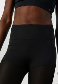 Close-up van een persoon in hoge zwarte sportlegging en een zwarte sporttop die de buik en bovenbenen toont.