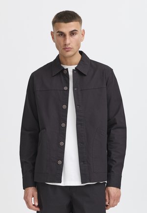 Mand med kort mørkt hår iført en sort knapjakke over en hvid skjorte, stående mod en ensfarvet hvid baggrund.