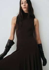 Braunes ärmelloses Kleid mit hohem Kragen, fließendem Rock und glatter Textur. Kombiniert mit enganliegenden schwarzen Lederhandschuhen.