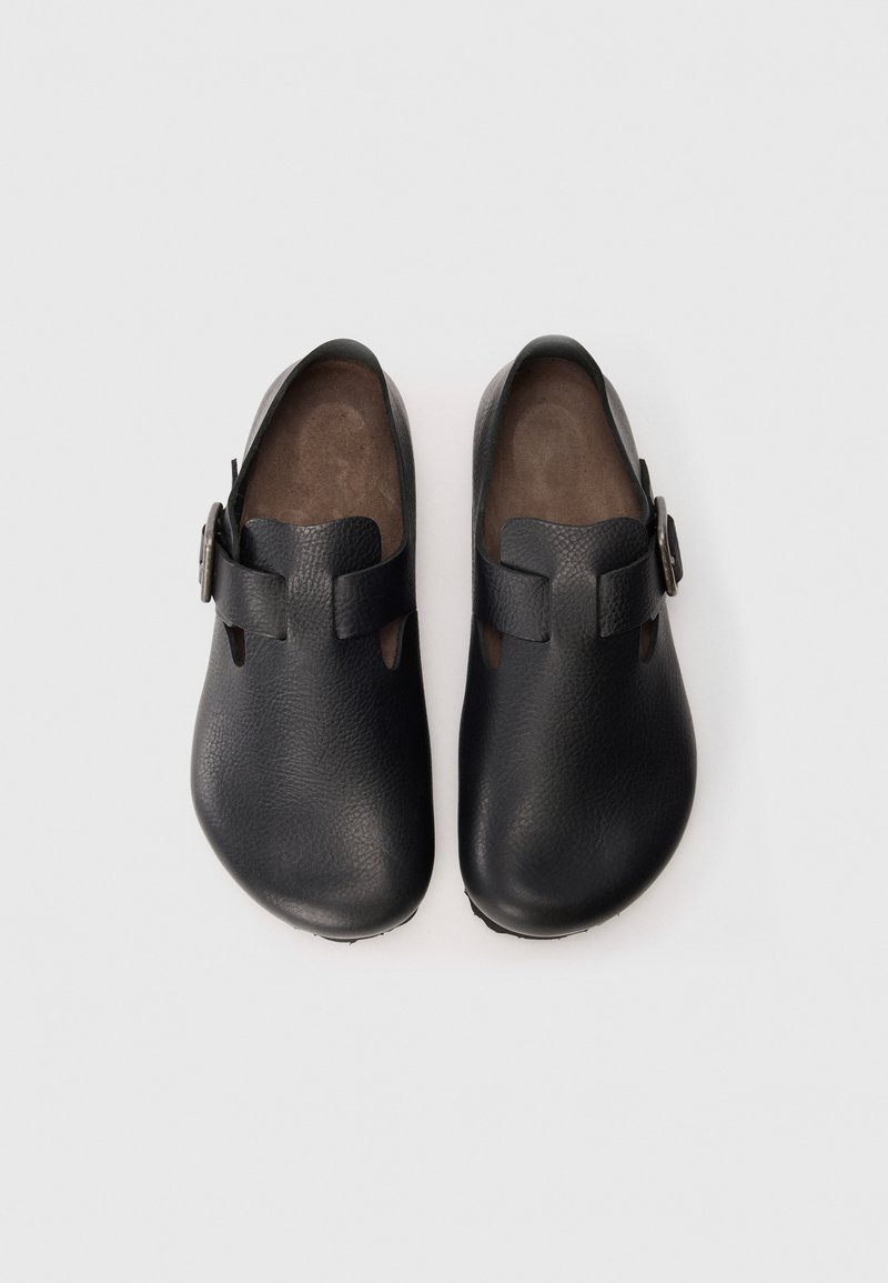 Black leather clogs with an open design, featuring cutouts and a metallic buckle accent. Smooth texture with a rounded toe shape.
