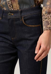 Jeans de denim azul oscuro con costuras en contraste, remaches decorativos en tono dorado a lo largo de los bordes de los bolsillos y un diseño clásico de cinco bolsillos.