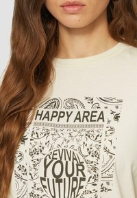 Lichtbeige katoenen t-shirt met grafische print, featuring "HAPPY AREA" en "REVIVAL YOUR FUTURE" in vetgedrukte zwarte tekst, omringd door sierlijke patronen.