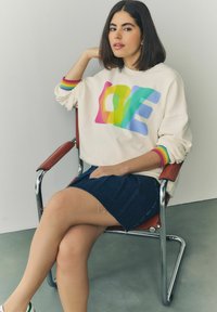 Krémová mikina s farebným grafikou "LOVE", rebrovými dúhovými manžetami, v kombinácii s tmavými denimovými sukňami. Modelka sediaca na chrómovej stoličke.