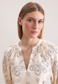 Lys cremekrage bluse med sorte paisley og blomster mønstre, rund halsudskæring og samlet detaljer på ærmerne. Glat stoftekstur.