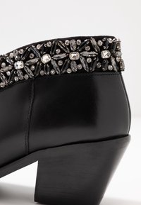 Botas de cuero negro hasta el tobillo con un tacón bajo, que cuentan con una banda decorativa de adornos de cristal negro y transparente a lo largo del borde superior.