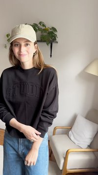 Crna dukserica s utisnutim natpisom "STUDIO", opuštenog kroja, okruglog izreza, u kombinaciji sa svijetloplavim trapericama, nosi se uz bež kapu.
