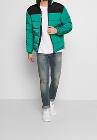 Grön puffjacka med svarta axlar, horisontell söm, framsida med dragkedja och elastiska ärmslut, som kombineras med blå jeans och vita sneakers.