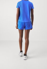 Persona vestida con una camiseta de manga corta azul brillante y pantalones cortos a juego, con zapatillas deportivas blancas y negras, de pie frente a un fondo sencillo.