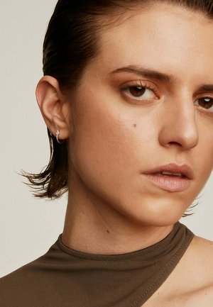 Arete de aro plateado en una persona con cabello corto y oscuro, que lleva una blusa marrón ajustada. La piel tiene una textura suave y luce un maquillaje natural.
