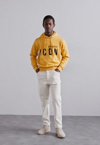 Felpa gialla con testo nero "ICON" e "DSQUARED2," abbinata a jeans beige chiaro e sneaker beige su uno sfondo semplice.