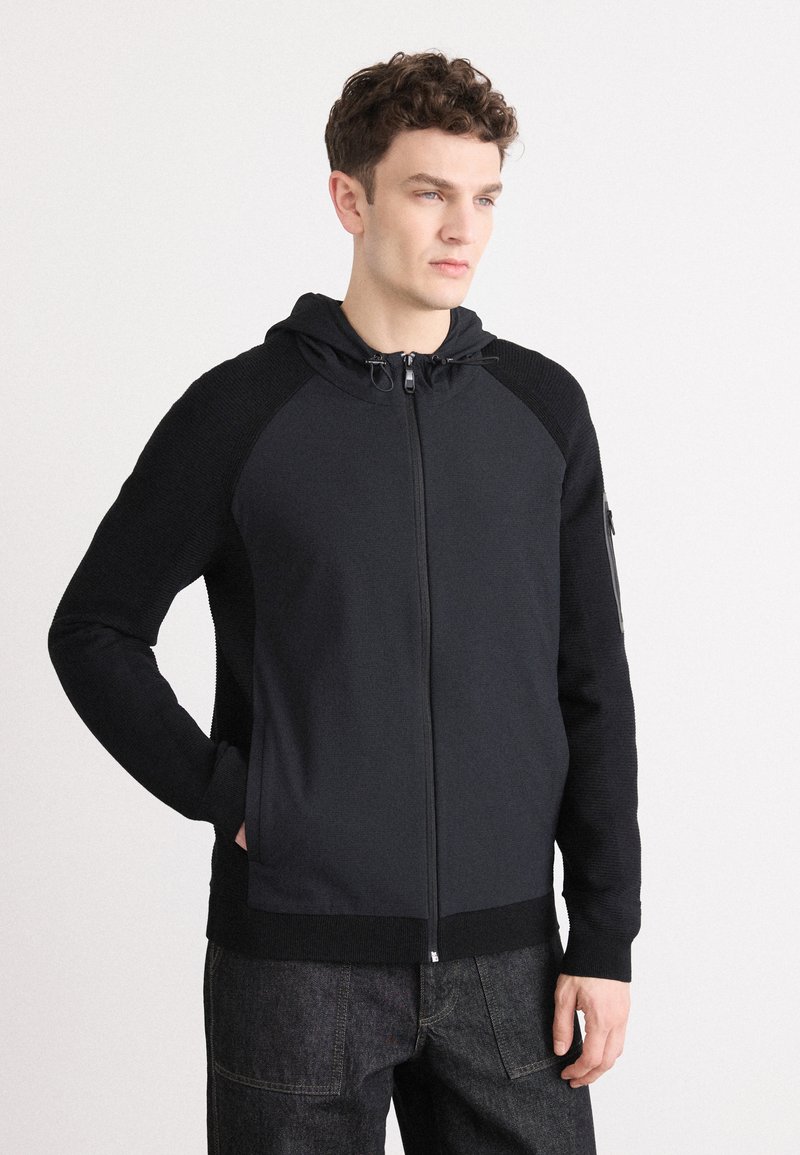 Moška zip-up kapuca v temno sivi barvi z teksturiranimi rokavi, nastavljivo kapuco in stranskimi žepi. Ima gladko sprednjo stran in rebraste manšete.