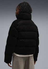 Sort corduroy pufferjakke med høj krave, der har quiltede sektioner og en cropped design. Bagfra ses den lette tekstur og den rundede form.