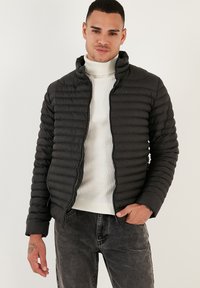 Giacca a vento nera con un design trapuntato, colletto alto e chiusura frontale con zip, indossata su un dolcevita bianco a coste. Presenta una vestibilità slim.