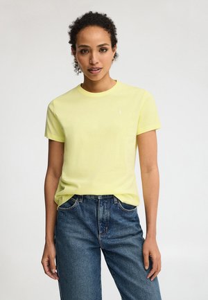 Kobieta o kręconych włosach ubrana w jasnoszarą żółtą koszulkę z krótkim rękawem i niebieskie dżinsy, stojąca na jednolitym jasnym tle.