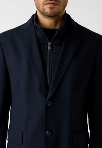 Marineblauwe overjas met gestructureerde revers, verborgen ritskraag en een dubbele knoopsluiting. Bevat een borstzak en zijzakken.