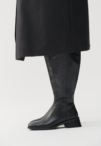 Botas de cuero negras hasta la rodilla con punta cuadrada y tacón bloque, con una textura suave y un detalle de costura minimalista.