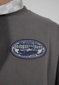 Chemise gris foncé avec un écusson brodé représentant une voiture vintage, un globe et le texte "PROHIBITED RACING CLUB" en bleu et blanc.