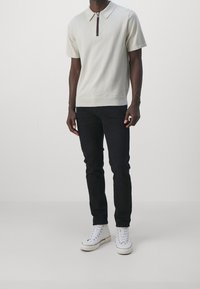 Světlý béžový polo tričko s krátkým rukávem a zipovým límcem, doplněné barevným pruhem. V kombinaci s úzkými černými džínami a bílými vysokými teniskami.