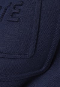 Κοντινή λήψη σκούρου μπλε υφάσματος με υφή, με ανάγλυφα ραμμένα γράμματα που αποτελούν μέρος σχεδίου ή λογοτύπου.