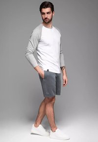 Šedé tričko s dlouhým rukávem raglánového stylu v kombinaci se šedými šortkami. Modelka má na sobě bílé tenisky, čímž prezentuje ležérní, moderní outfit.