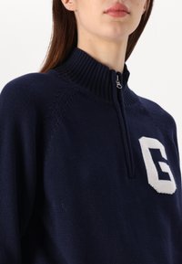 Mörkblå halvdraget tröja i stickad tyg med ribbad krage. Har en kontrasterande vit "G"-logotyp på vänster bröst. Strukturerad detaljering på axlarna.