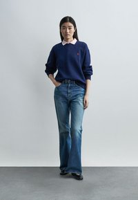 Νεαρή γυναίκα που φοράει μπλε πουλόβερ με ροζ γιακά, μπλε τζιν καμπάνα και μαύρα παπούτσια, στέκεται μπροστά σε απλό ανοιχτό γκρι φόντο.