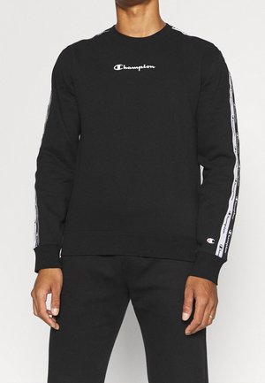 Homme portant un sweat Champion noir avec un logo blanc centré sur la poitrine et des bandes répétées avec le logo le long des deux manches.