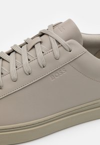 Beige lädersneakers med intrikata snördetaljer, präglad logotyp och en strukturerad gummisula. Minimalistisk design, smidig yta och subtila stygn.